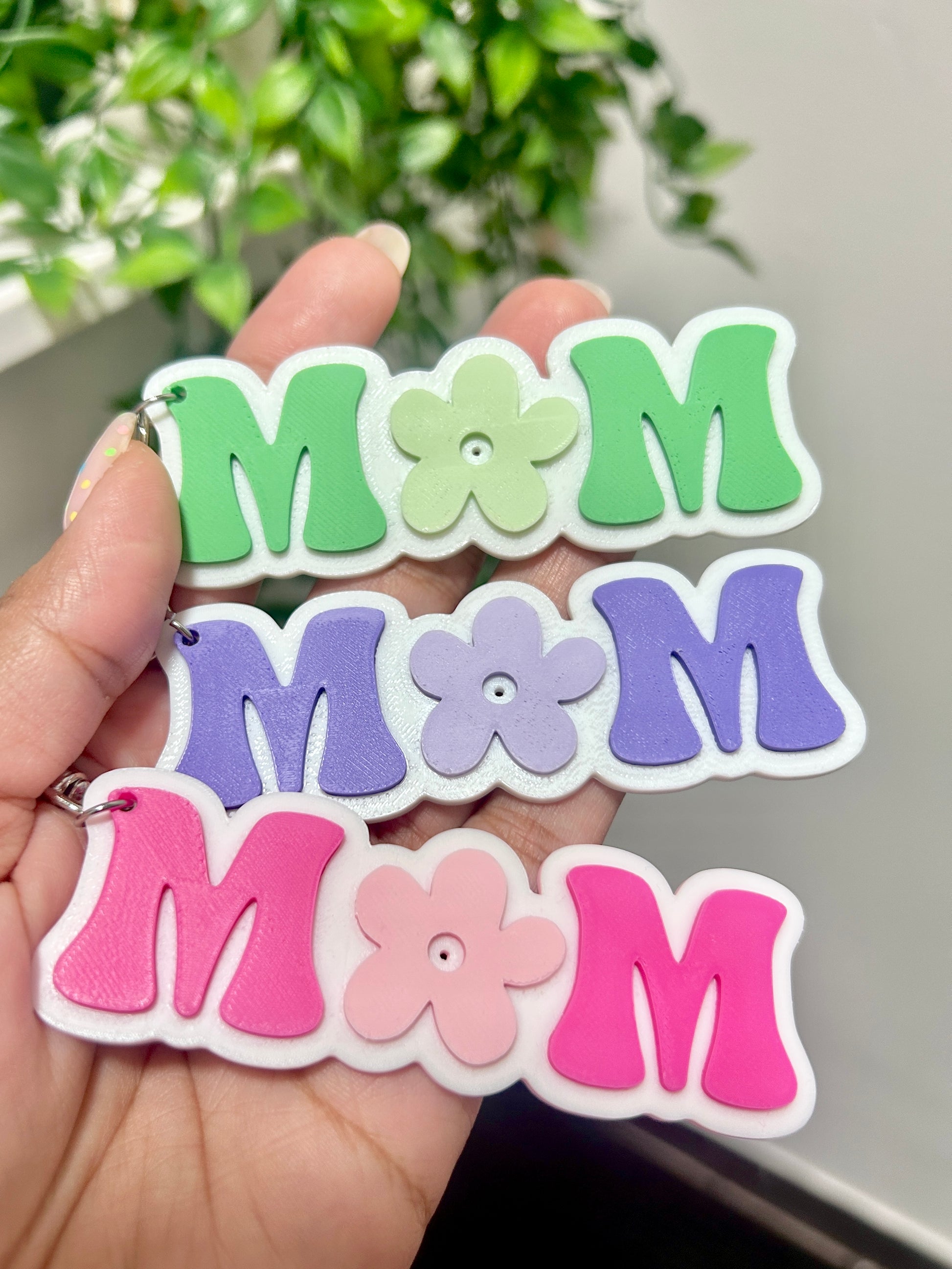 Colorful 'MOM' keychain held in a hand with a plant in the background.