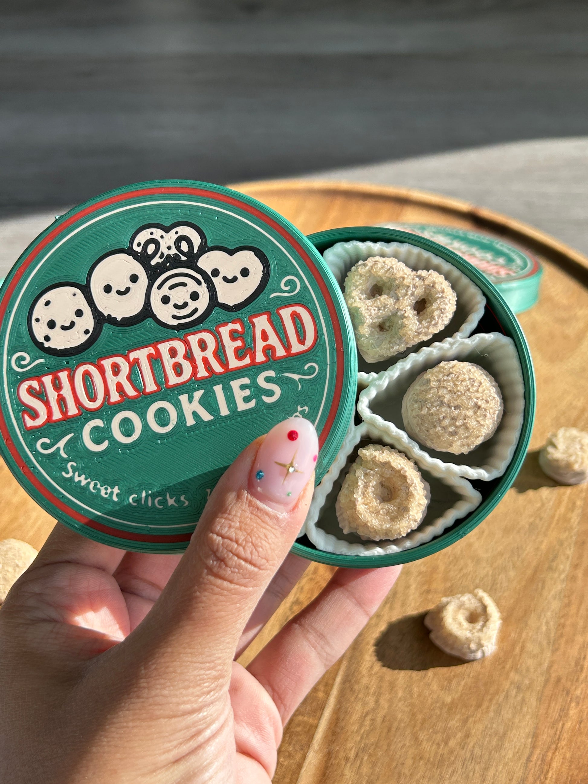 Hand holding a container of shortbread cookies with a decorative lid on a wooden surface.