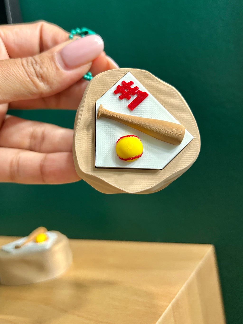 Hand holding 3D printed mound of dirt with home plate and softball laying next to bat next to #1 text as a fidget clicker keychain.
