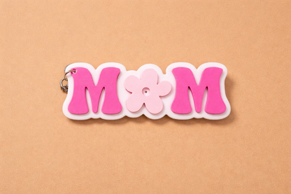 Pink and white 'MOM' keychain on a beige background.
