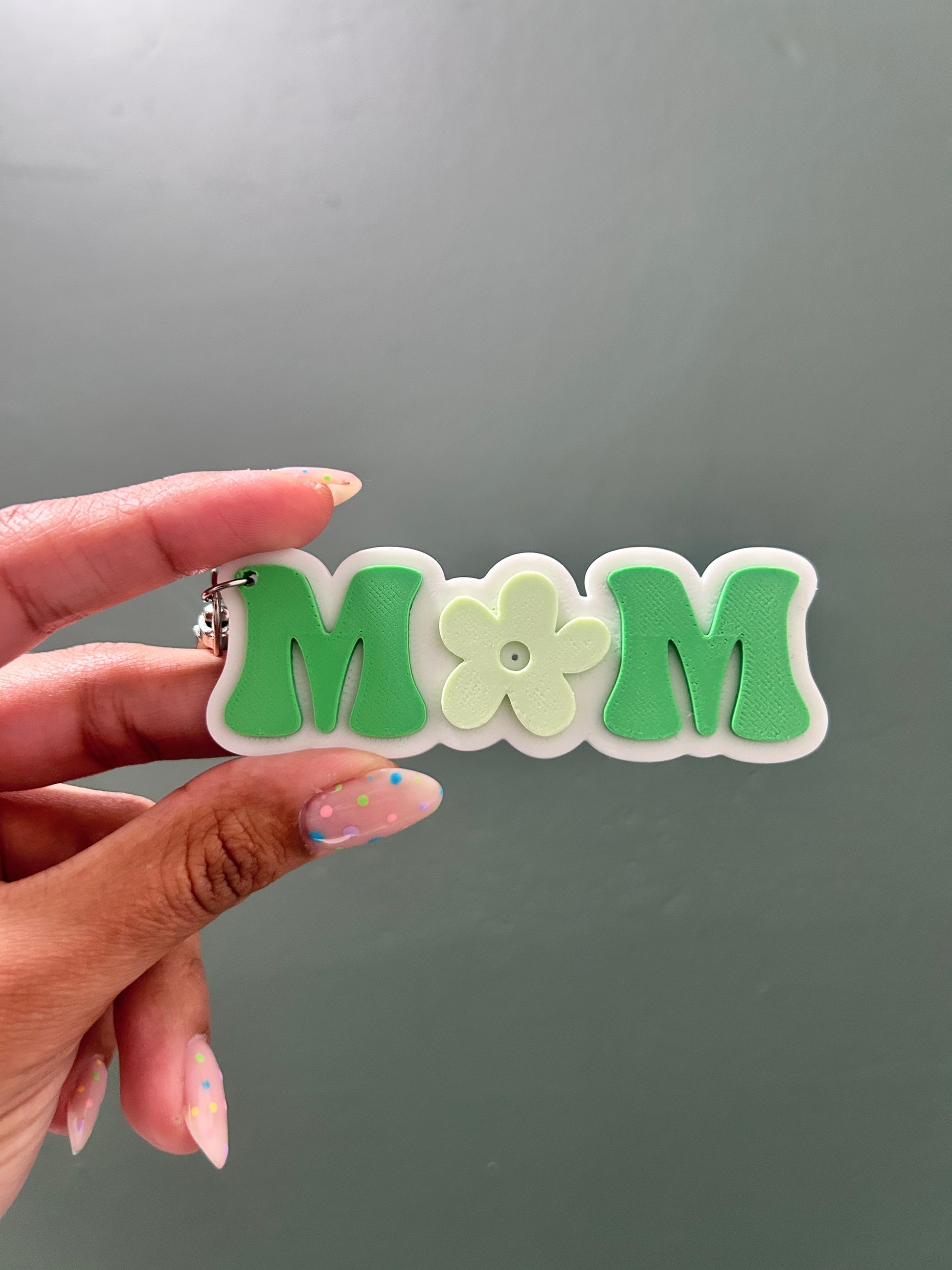 Hand holding a green and white decorative keychain with 'MOM' and a flower design against a gray background.