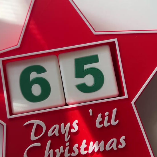 Video pans around a red, star-shaped Christmas countdown sign with '65 Days 'til Christmas' laying on its back on a white shelf.
