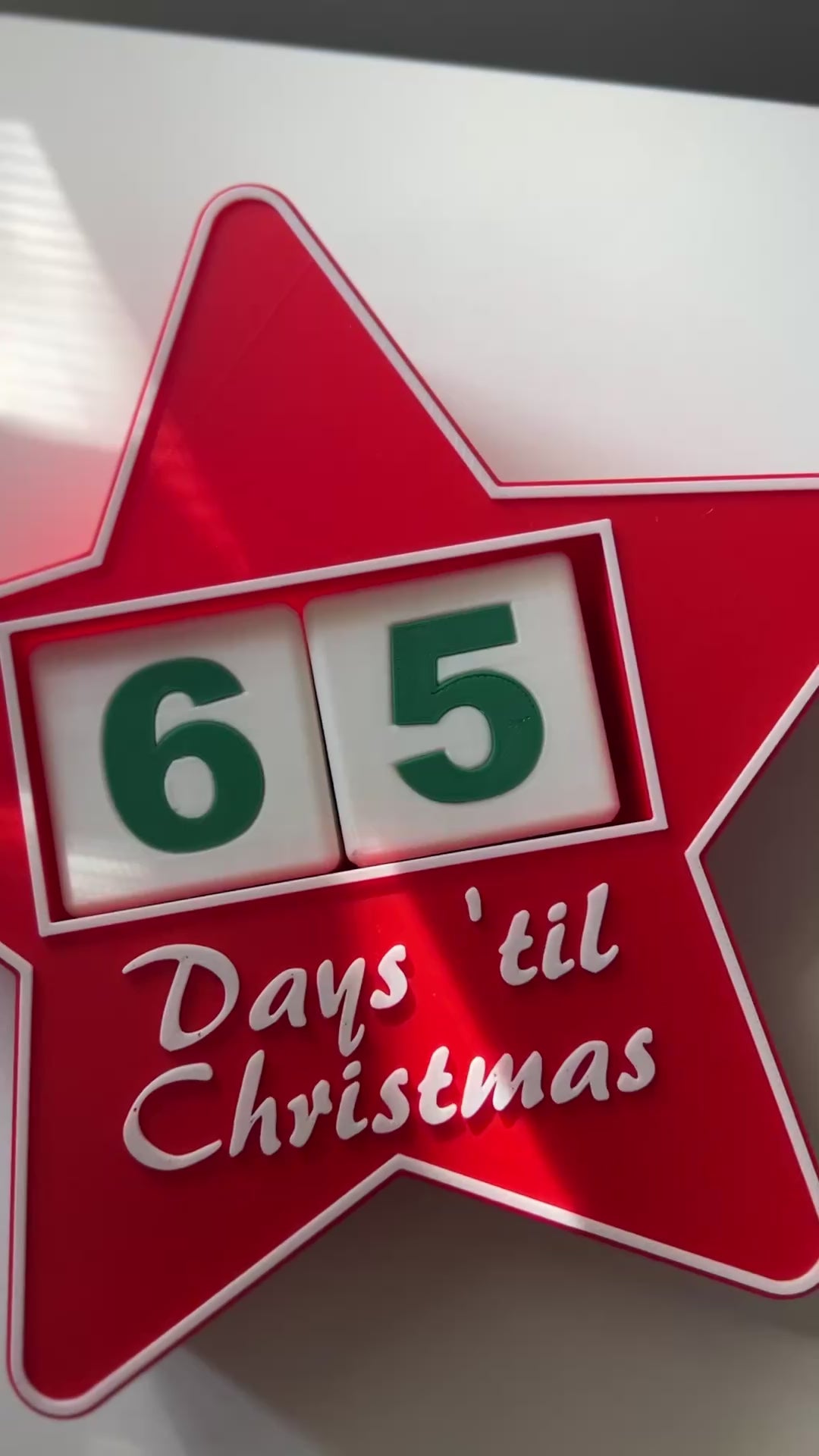 Video pans around a red, star-shaped Christmas countdown sign with '65 Days 'til Christmas' laying on its back on a white shelf.