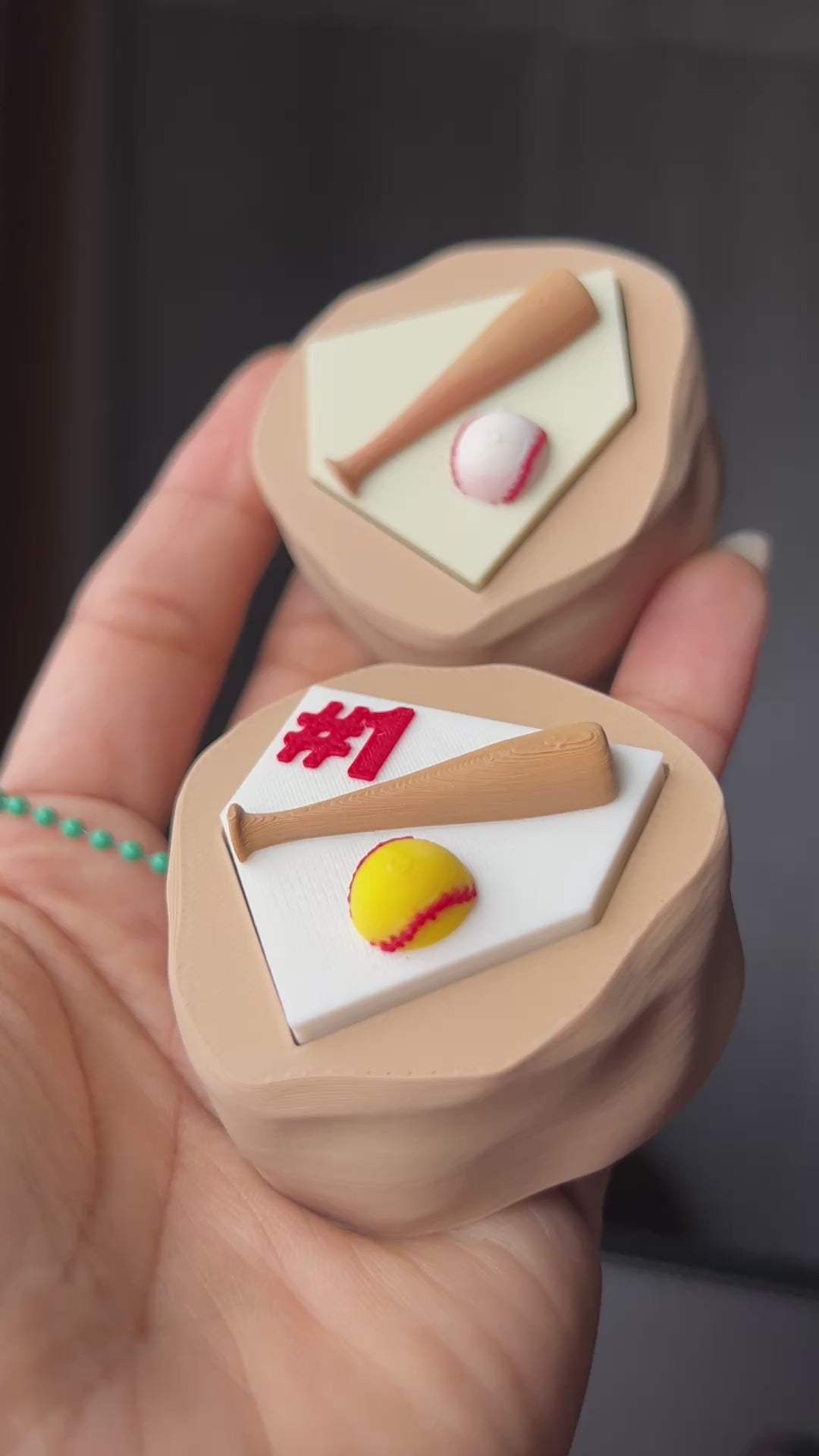 Hand holding one baseball and one softball fidget clicker to show off the custom in-house design by PLAground Shop.