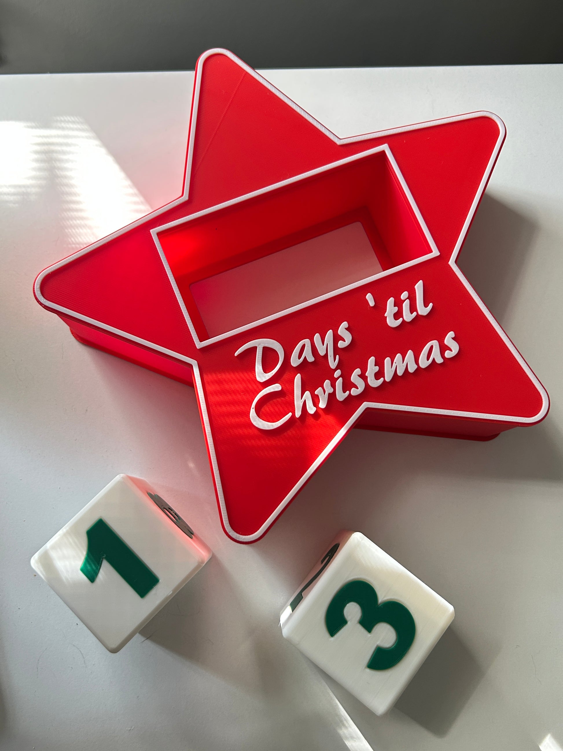 Red star-shaped container with 'Days 'til Christmas' text, surrounded by countdown blocks revealing a number one face and a number three face on a white surface.