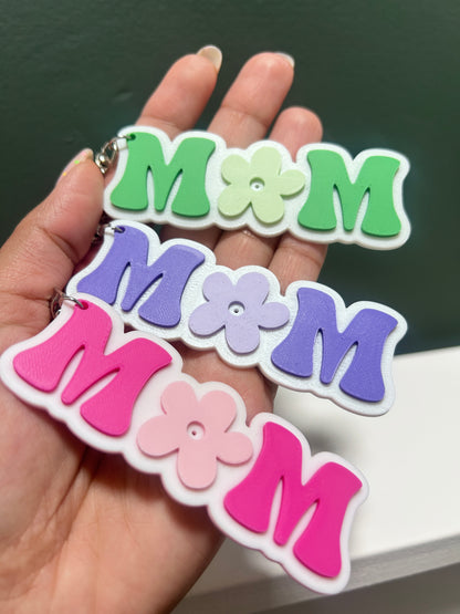 Colorful 'mom' keychains held in a hand against a neutral background.